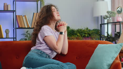 Young-woman-girl-praying-sincerely-with-folded-arms-asking-God-for-help-begging-apology-on-home-sofa