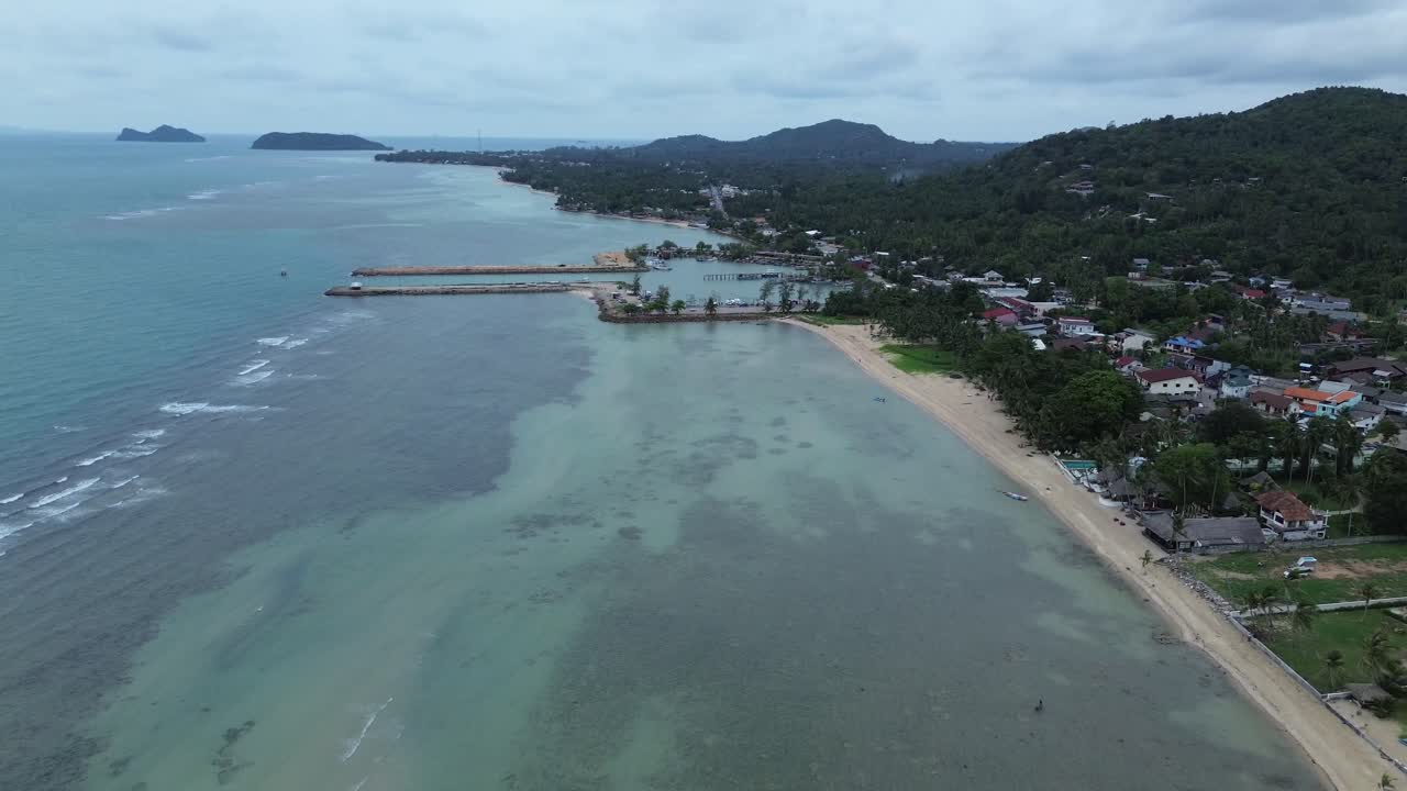 Aerial drone footage showing Baan Tai Pier and the surrounding coastal town on Koh Phangan island, Surat Thani, Thailand, with tropical hills, beach shoreline, and blue sky