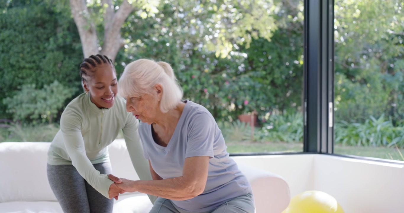 African American carer indicating seat reaching hand assisting senior woman onto sofa by windows