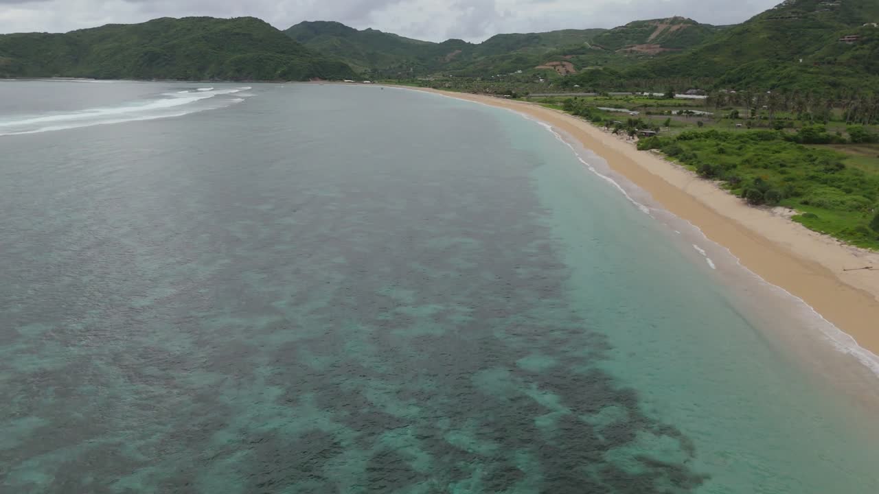 Drone-luftoptagelse langs den kurvede kystlinje ved Pantai Lancing, også kendt som Tampah Beach, nær Kuta Lombok. Turkis vand, koralrevsmønstre, blide bølger og grønne bakker under overskyet regnvejr