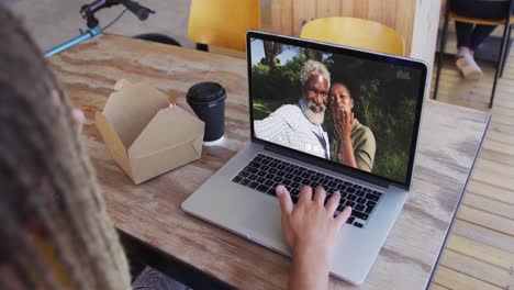 Woman-having-a-snack-while-having-a-video-call-on-laptop-at-a-cafe