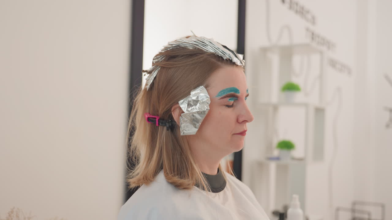 Hvid kvinde i salon med folie og turkis øjenbrynsmakeup sidder ned, mens klient får highlights med folie. Stylist forbereder sektioner, modelpose i redaktionel stil, rolig profil og trekvartvisning, neutral