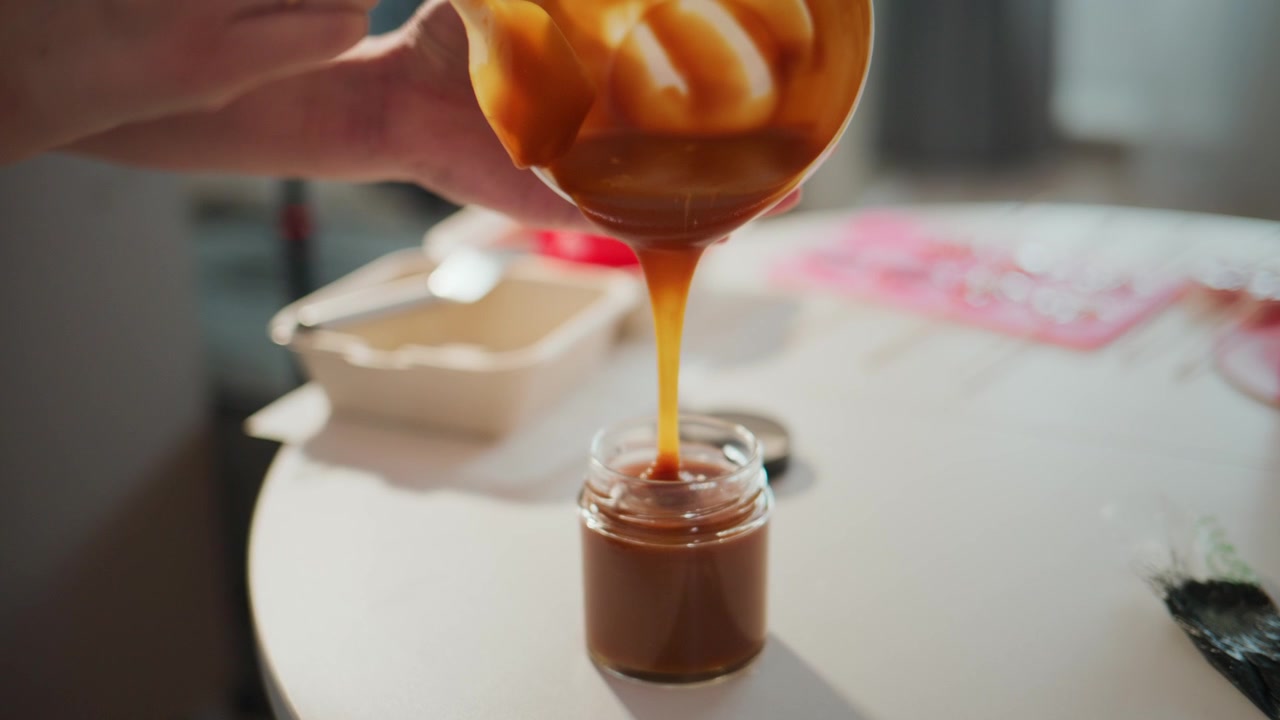 Mani versano una densa salsa al caramello in un barattolo. Lo sciroppo dorato scorre lentamente, creando ricche consistenze. Un tavolo bianco contiene utensili e contenitori. La luce brillante evidenzia il dolce processo. Perfetto per blog di cucina
