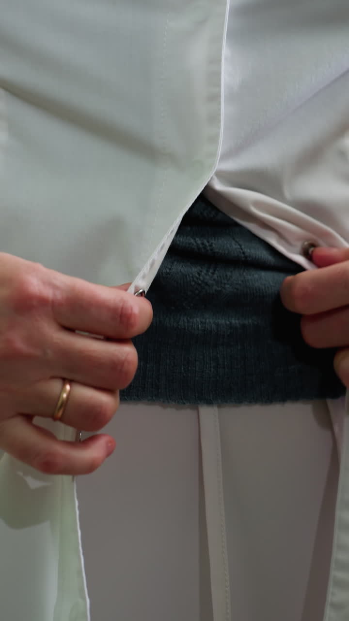 흰색 코트를 잠그는 여성 간호사, 회색 스웨터 위에 단추를 채우는 손, 손가락의 결혼 반지, 집중된 차분한 표정, 복도 배경은 클리닉을 암시하며, 클로즈업 샷은 직물을 강조합니다.