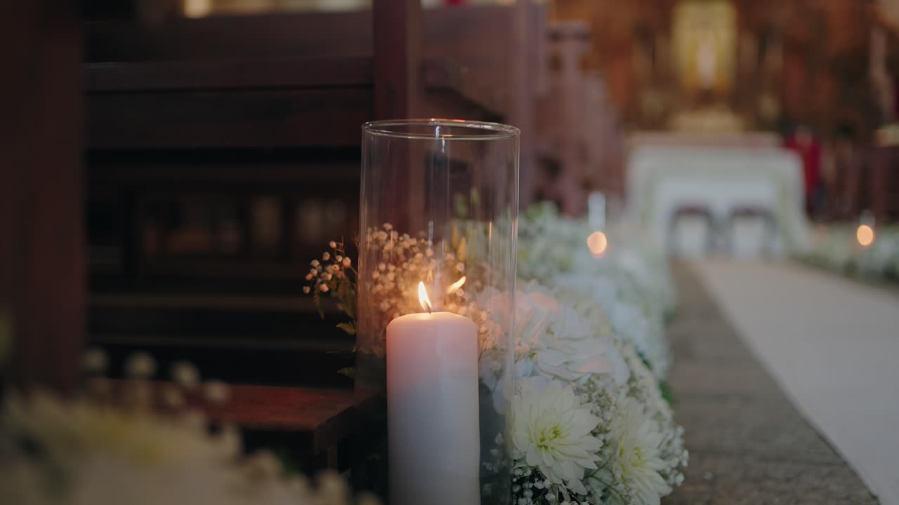 Candela accesa in un vaso di vetro con decorazioni floreali bianche lungo la navata della chiesa.
