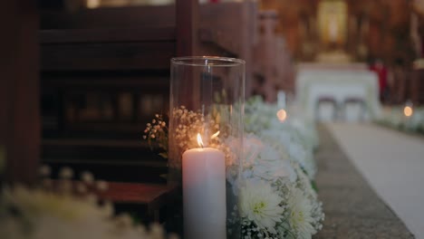 lit candle in glass vase with white floral decor along church aisle
