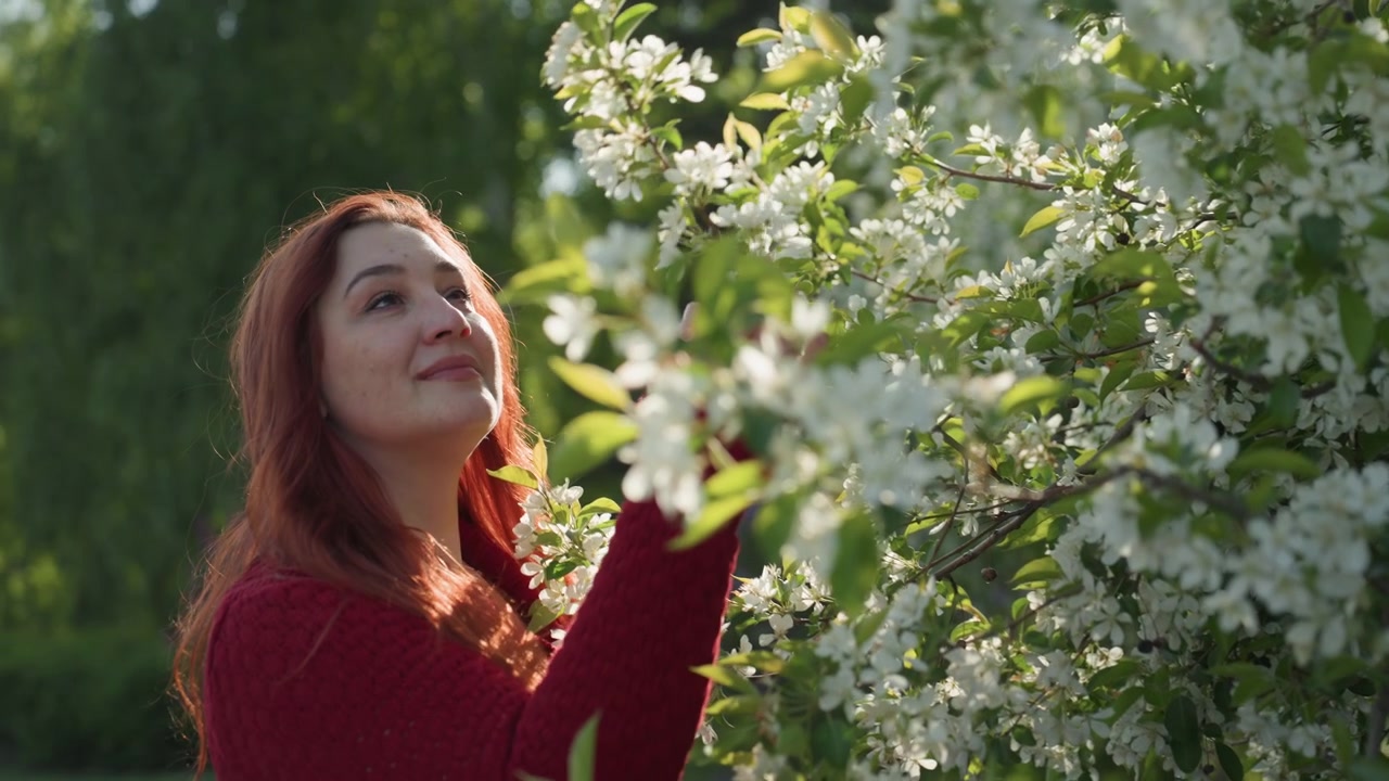 Primo piano di donna con lunghi capelli rossi che indossa un maglione bordeaux, mentre tocca e annusa delicatamente fiori bianchi su un ramo d'albero all'aperto. La scena è caratterizzata da una luce naturale soffusa che filtra attraverso il verde.