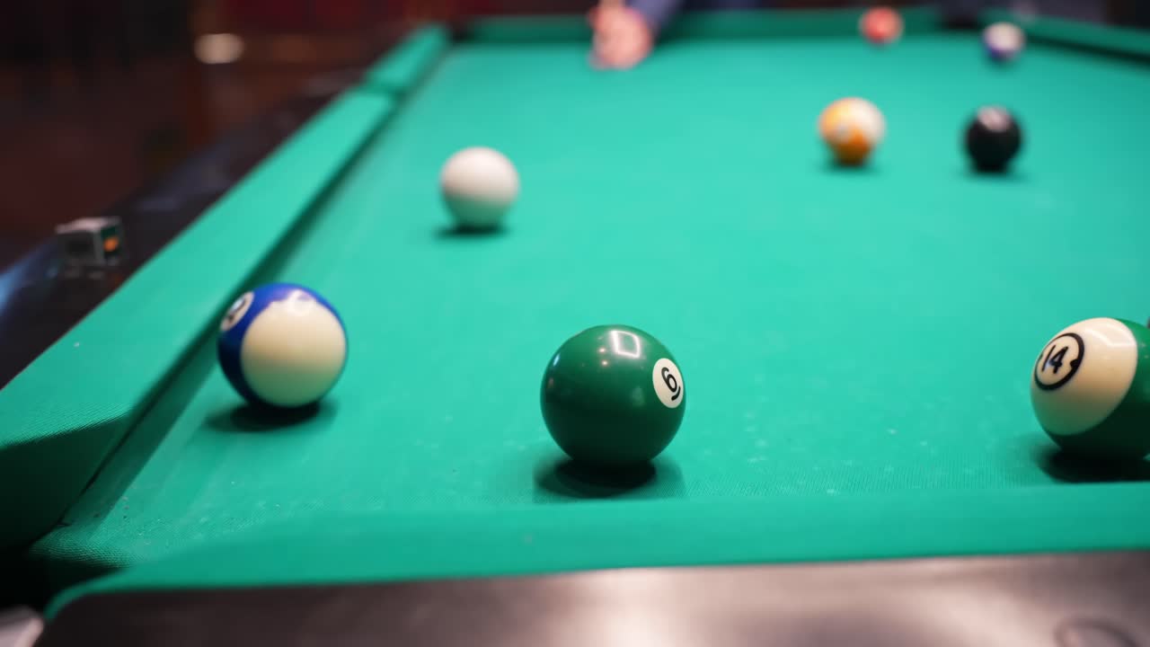 Close-up pool table as a blue ball drops into the corner pocket, cue ball and striped numbers on green felt with shallow depth of field in a bar game setting