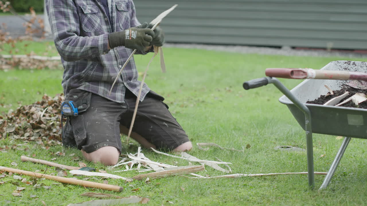 A man kneels on a lawn, splitting wooden stakes from branches, with a wheelbarrow nearby.