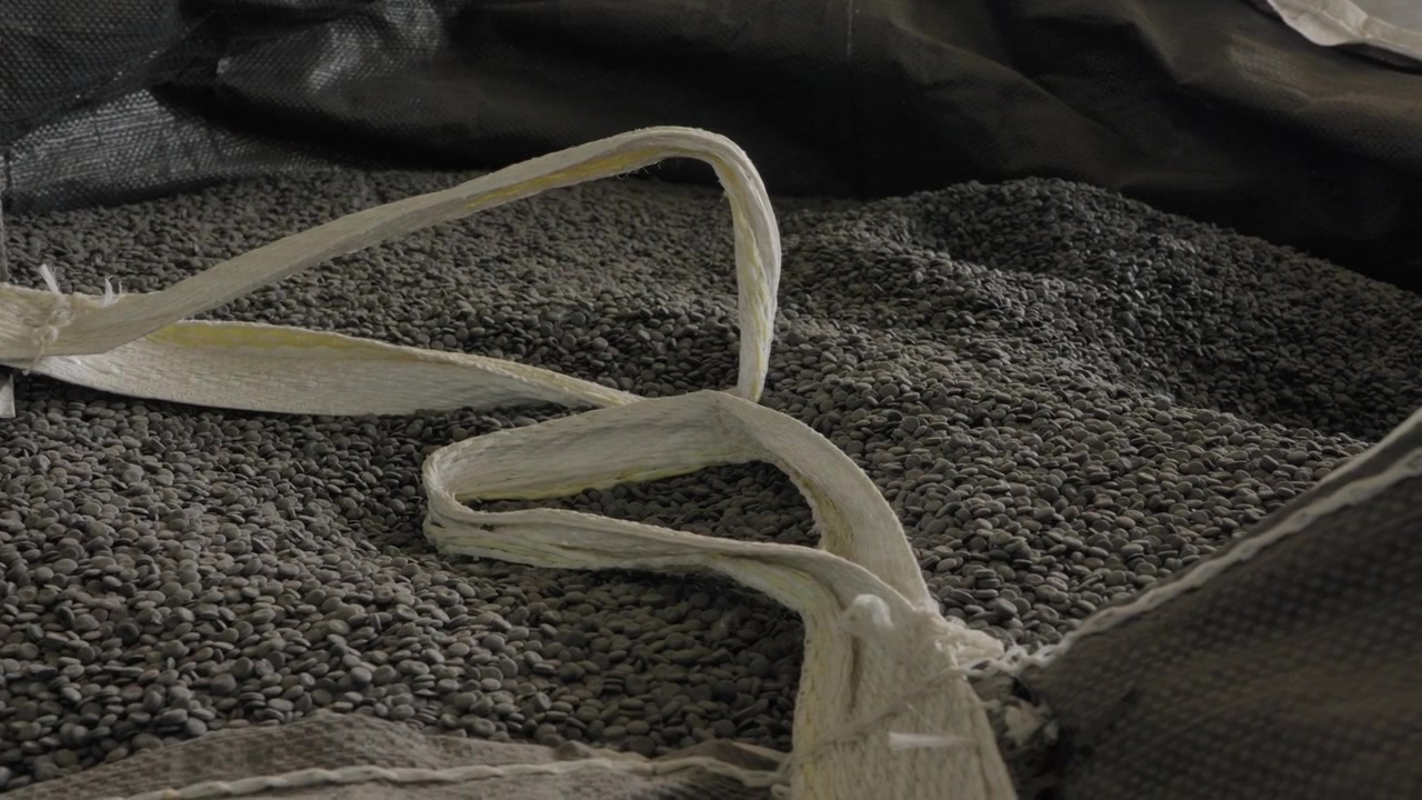 Hvide, iturevne stropper fra bigbags ligger hen over en tæt, mørkegrå bunke genanvendte plastgranulater på en genbrugsfabrik