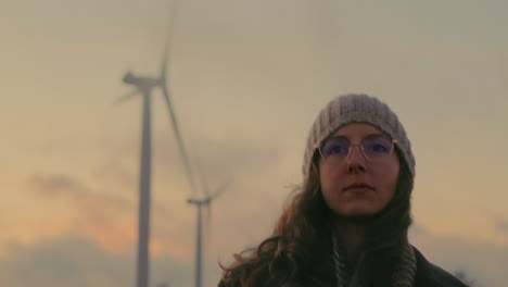 in cinematic portugal, a young blonde woman with glasses walks near wind turbines during a breathtaking sunset. she enjoys the beautiful scenery while promoting sustainable energy and nature therapy.