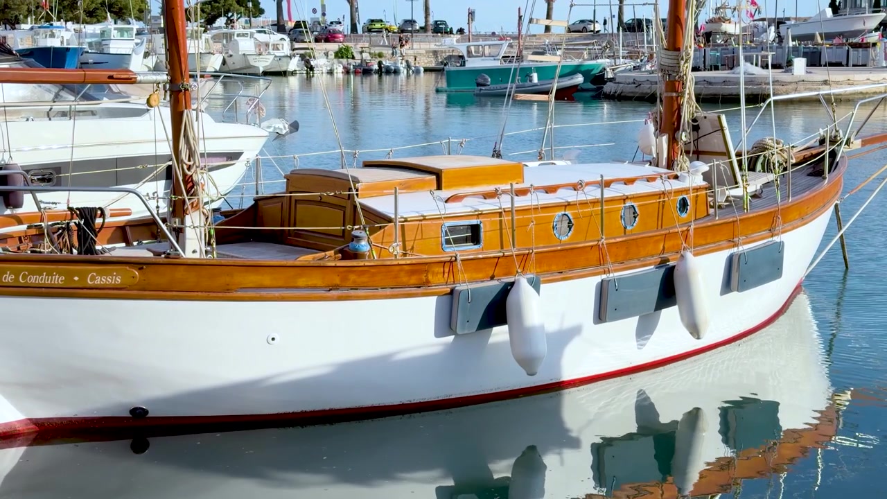 Una barca a vela d'epoca in legno galleggia su acqua turchese in un soleggiato porto turistico mediterraneo