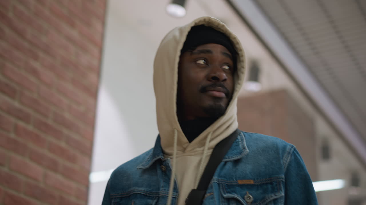handsome black man wearing hoodie under denim jacket strolling through modern shopping center, glancing upward at overhead lighting and displays, urban casual style portrait with soft ambient glow