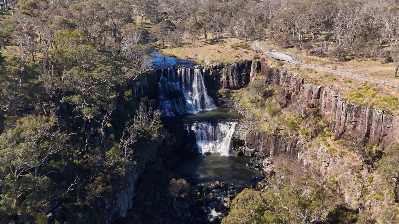 Drone camera glides toward Ebor Falls, revealing cascading water, rocky cliffs, and surrounding dry woodland under bright natural daylight