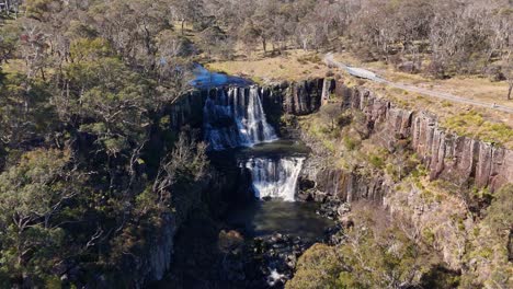 drone camera glides toward ebor falls, revealing cascading water, rocky cliffs, and surrounding dry woodland under bright natural daylight