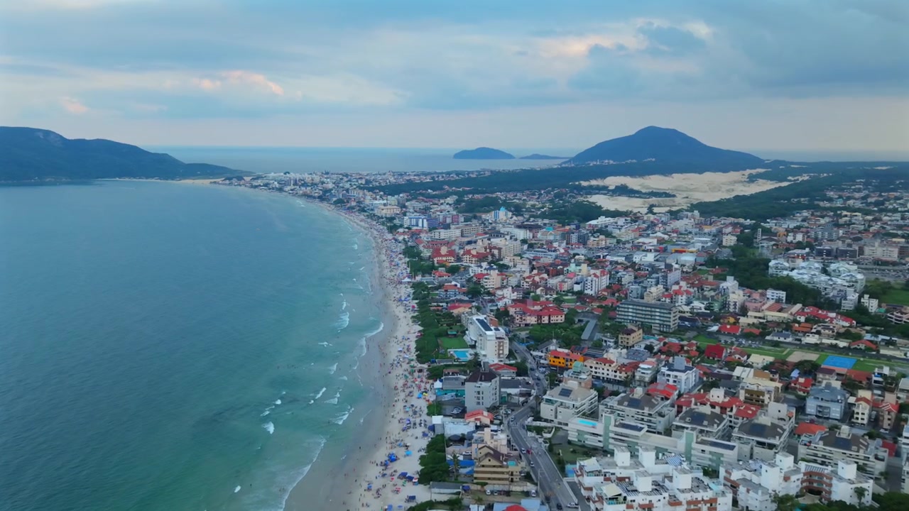 Majestosa tomada aérea de estabelecimento capturando a vasta escala do desenvolvimento urbano costeiro no Brasil, perfeitamente aninhado entre o oceano, montanhas e dunas de areia.