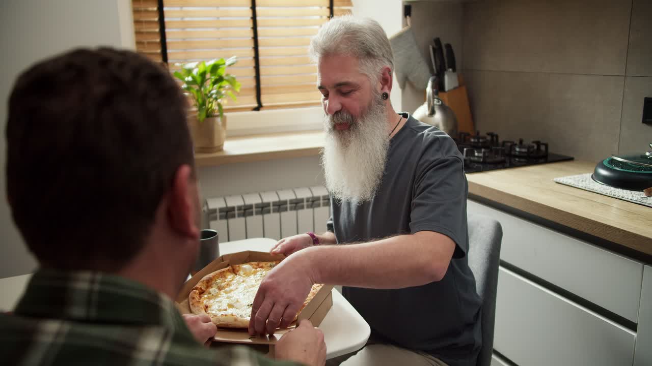 hombre feliz con el color del cabello gris y la barba blanca exuberante con una camiseta gris come pizza con su novio moreno en una camisa a cuadros en la cocina de una caja de entrega de cartón