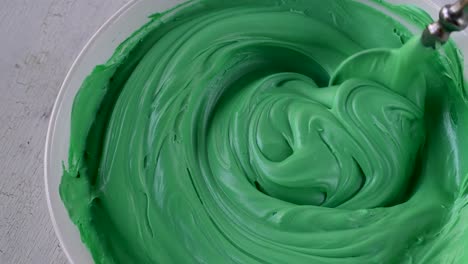 mixing light green buttercream icing in bowl with spoon on white background top view