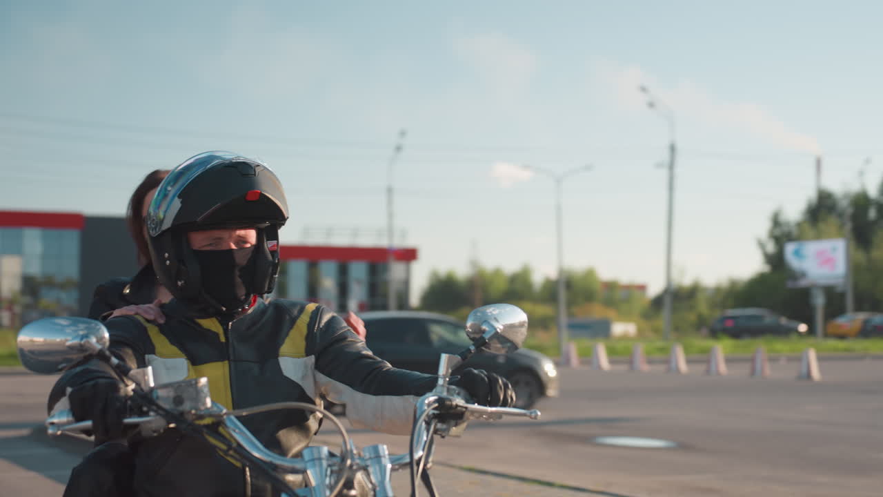 Young woman in leather approaches motorcycle beside helmeted rider, preparing to climb onboard before departure from sunlit urban parking lot, mirrors gleaming, traffic and cones in background