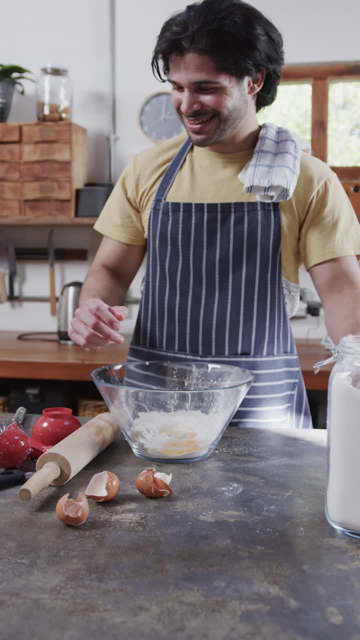 Vertical video of happy biracial man in apron preparing bread dough in kitchen, slow motion