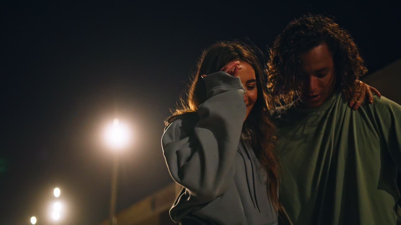 una pareja joven caminando por la calle de la noche en primer plano. un patinador feliz equilibrando un patineta.