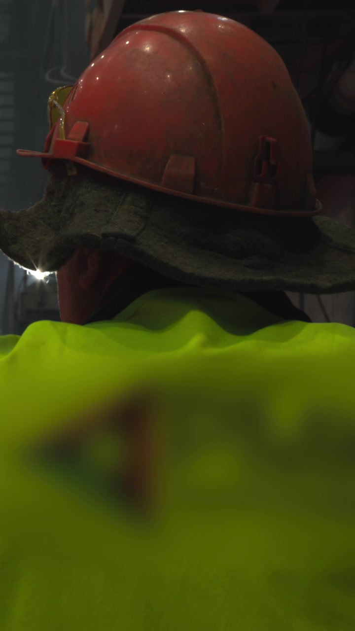 Operaio con gilet ad alta visibilità che osserva un forno incandescente, vista di spalle con casco e occhiali di protezione, ringhiera in primo piano.