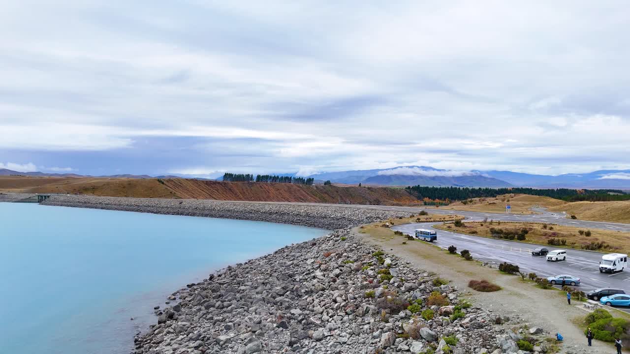Drone footage captures Lake Pukaki's tranquil waters and rocky shoreline under overcast skies, with parked vehicles along the road