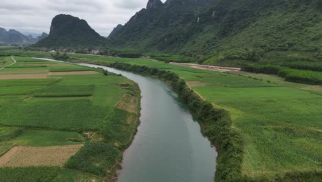 Aerial-Drone-Shot-Of-River-Passing-Through-Crop-Field-Near-Mountain-Rangers