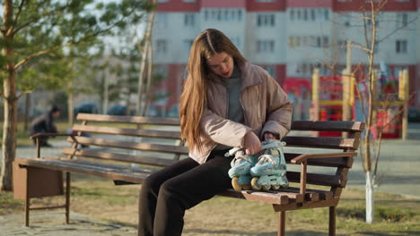 a girl wearing a peach jacket, grey inner shirt, and black trousers is sitting on a park bench with her rollerblades beside her. calm moment of rest during a sunny day in the park