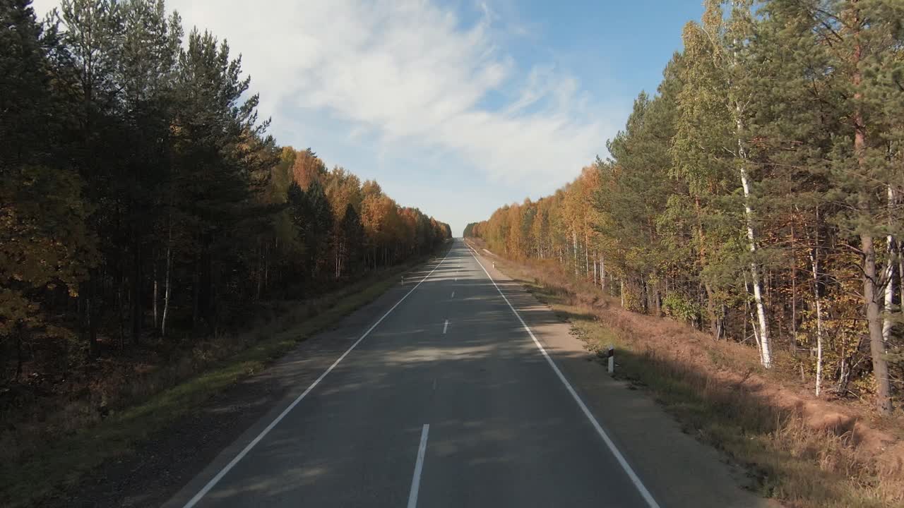 Filmato drone FPV che vola sopra una lunga strada asfaltata vuota che attraversa una colorata foresta autunnale sotto un cielo blu, concetto di viaggio e trasporto panoramico in campagna.