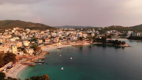 Drone-view-in-Albania-flying-over-blue-crystal-clear-water-on-sunset,-white-sand-beaches-and-hotels-with-red-light-in-Ksamil