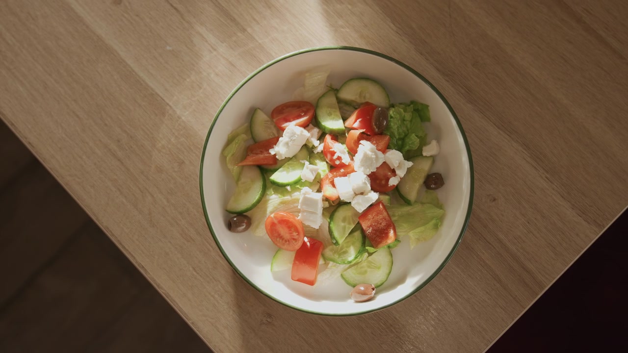 Close up of fresh salad with tomatoes cucumbers olives feta cheese in white bowl on wooden table. Salad ingredients like lettuce and red bell pepper are visible under soft lighting. Bowl