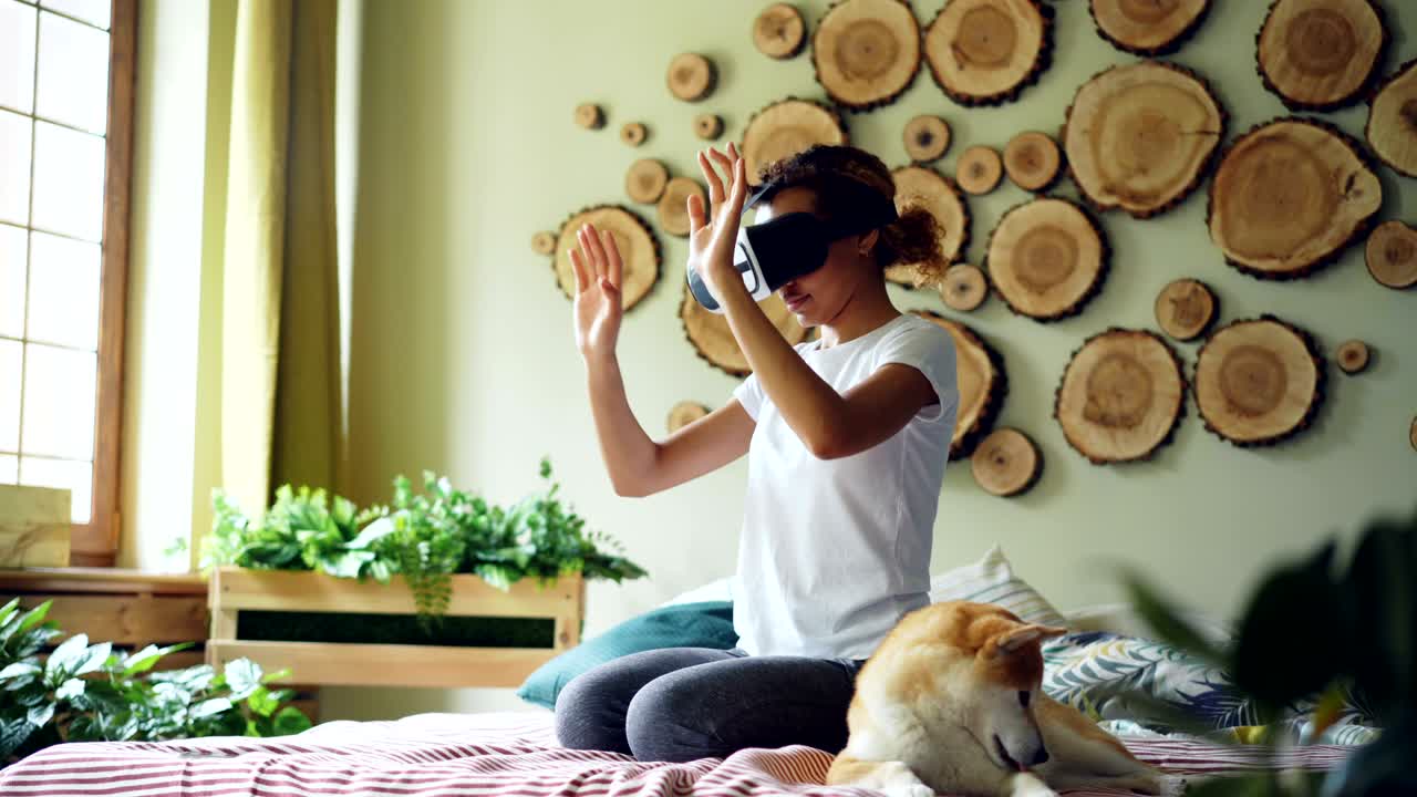 una chica afroamericana está usando gafas de realidad virtual sentada en la cama en casa, está moviendo las manos y la cabeza y sonriendo mientras su lindo perro está bostezando y lamendo su hocico.