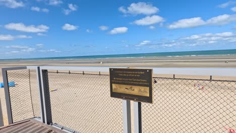 camera pans across metal sign on boardwalk, revealing sandy beach, blue sky, and sea