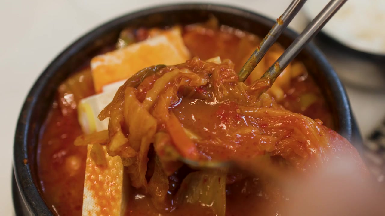 Tampilan jarak dekat sup kimchi tradisional Korea dengan tahu yang diaduk menggunakan peralatan logam dan sendok.