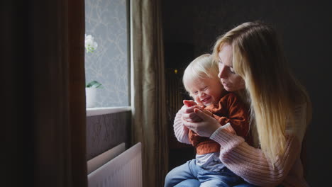 mother cuddling son trying to keep warm by radiator at home during cost of living energy crisis