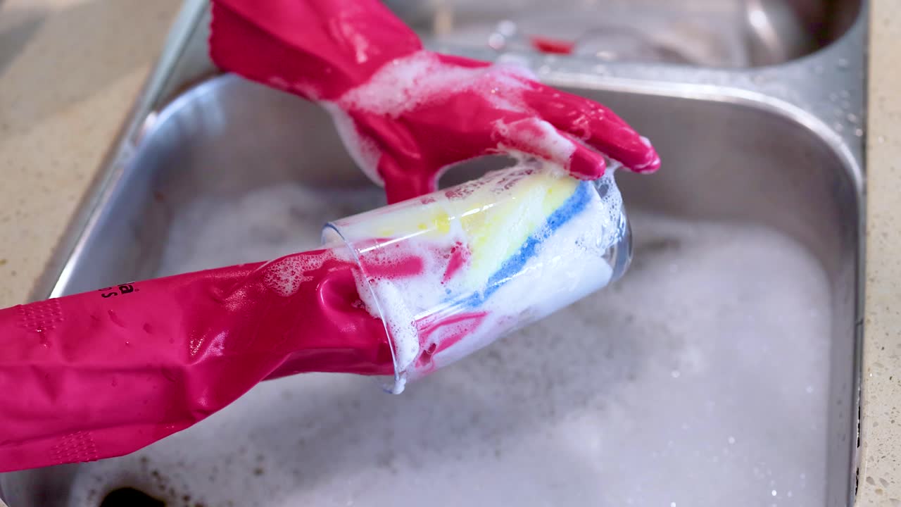 Hands in red gloves wash a glass with a sponge in a kitchen sink. Bright lighting highlights the cleaning process