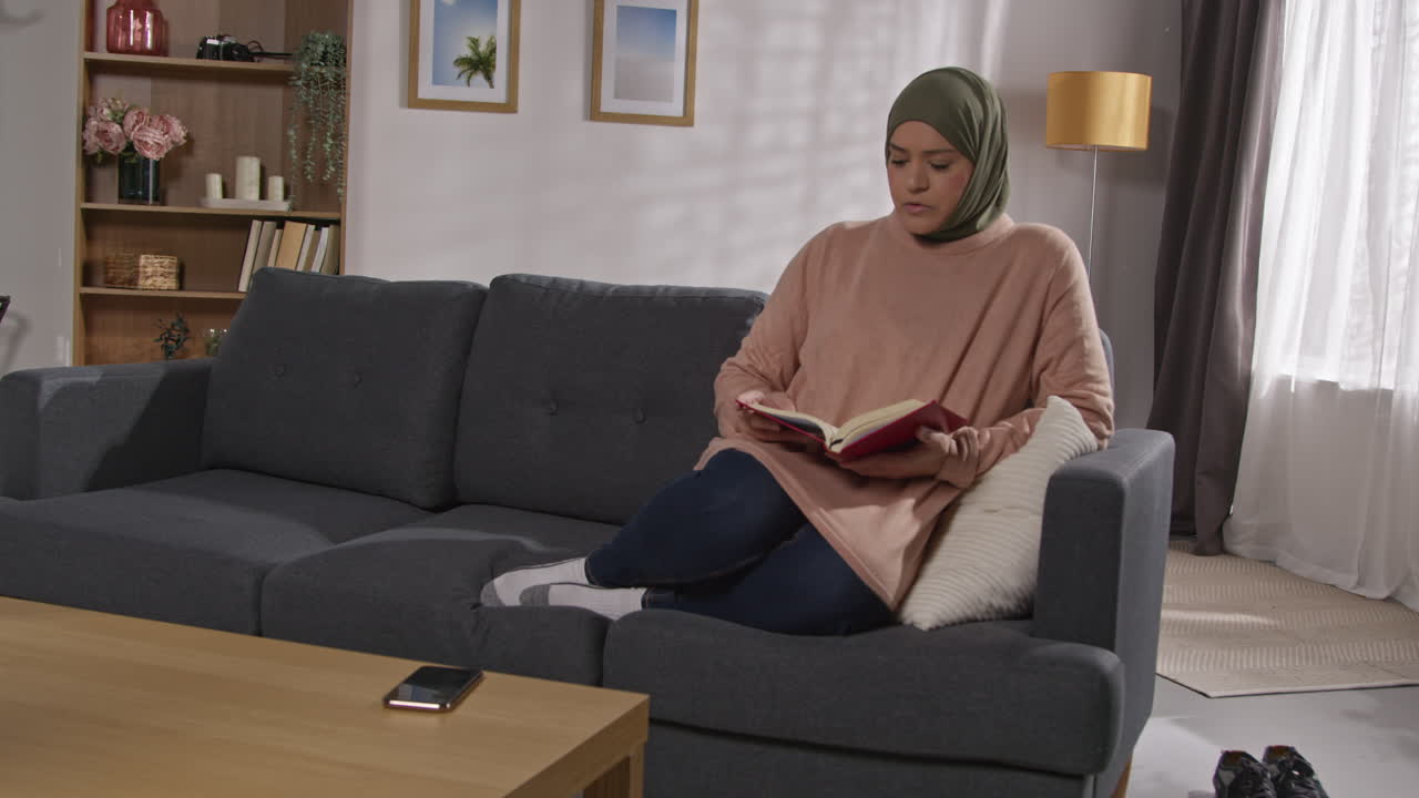Muslim Woman Wearing Hijab Sitting On Sofa At Home Reading Or Studying ...