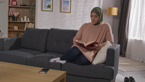 muslim woman wearing hijab sitting on sofa at home reading or studying the quran