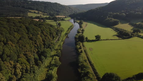 drone footage of the gentle flowing river wye on a summers day, with a clear blue sky, surrounded by green fields and a forested woodland valley in the background.
