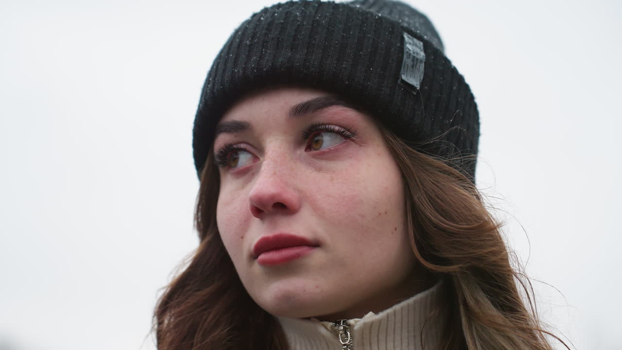 Primo piano di una donna con un berretto nero che cammina con i capelli al vento e uno sguardo pensieroso su uno sfondo cittadino sfocato durante una fredda e tranquilla giornata invernale con cielo nuvoloso e luce naturale soffusa