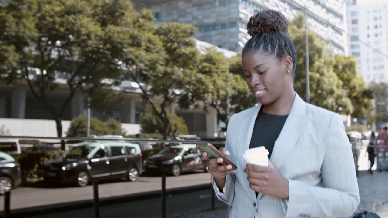 Dolly shot of a happy African-American businesswoman walking outside, holding coffee and using mobile phone