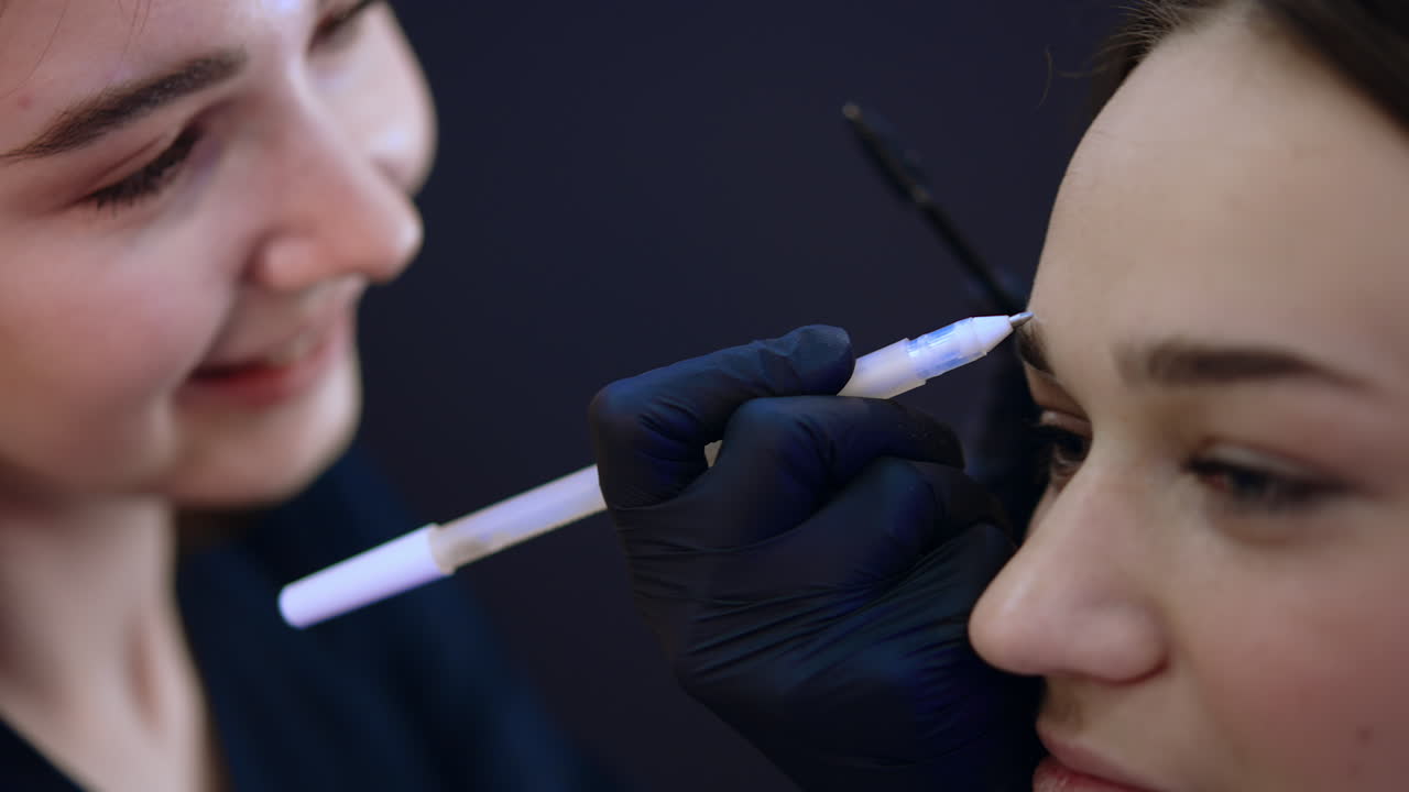 Caucasian female beautician drawing line above the eyebrow. Microblading, micropigmentation eyebrows work flow in a beauty salon. Extreme close up.