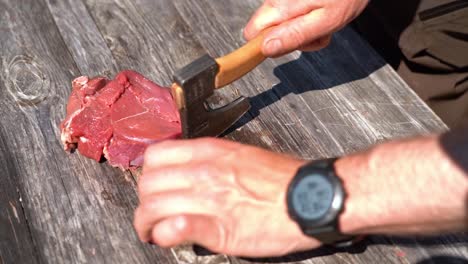 male hands using small hiking axe to cut filets from raw meat outdoor on wooden table - food preparation in nature