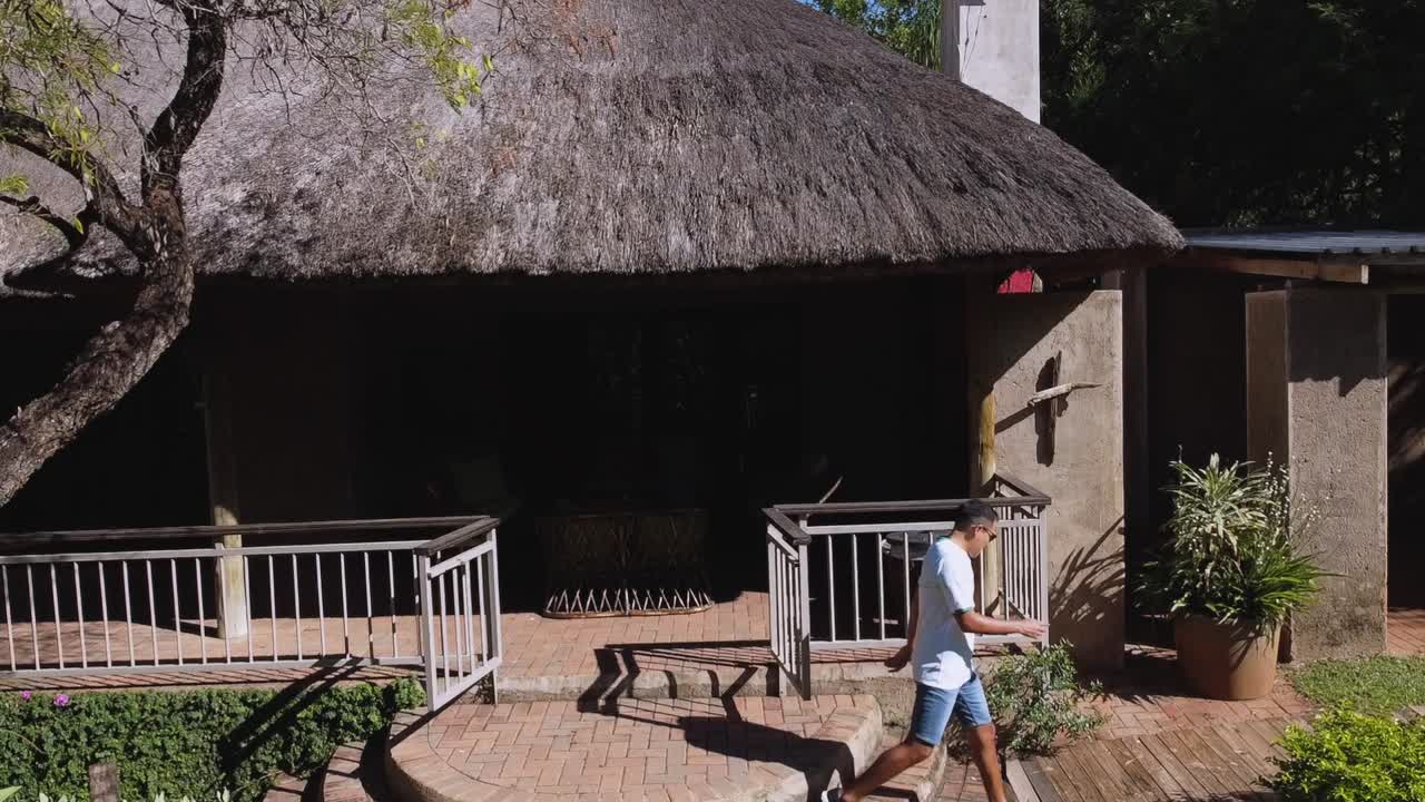 흰색 티셔츠와 파란색 반바지를 입은 한 남자가 화창한 날 남아프리카공화국 크루거 국립공원의 숙소 캠프에서 걸어 나오는 비디오