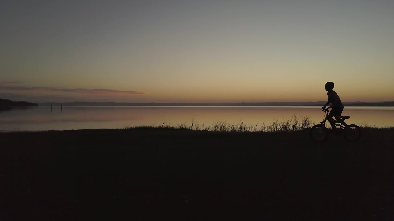 Silhouette of happy young boy riding his bike on shore next to beautiful calm lake reflecting vibrant colours during twilight hours- track and follow shot.