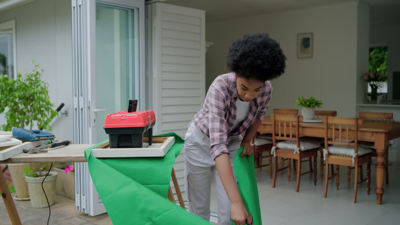 Vídeo vertical: Mulher de xadrez desdobrando tecido e aparando no pátio para arrumar com tesoura, com caixa de ferramentas.