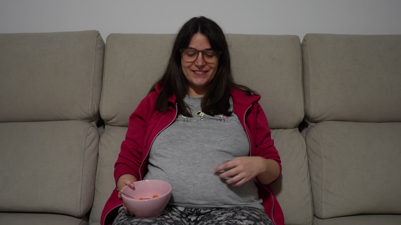 Pregnant woman smiling with bowl of fruit on sofa while feeling baby movements