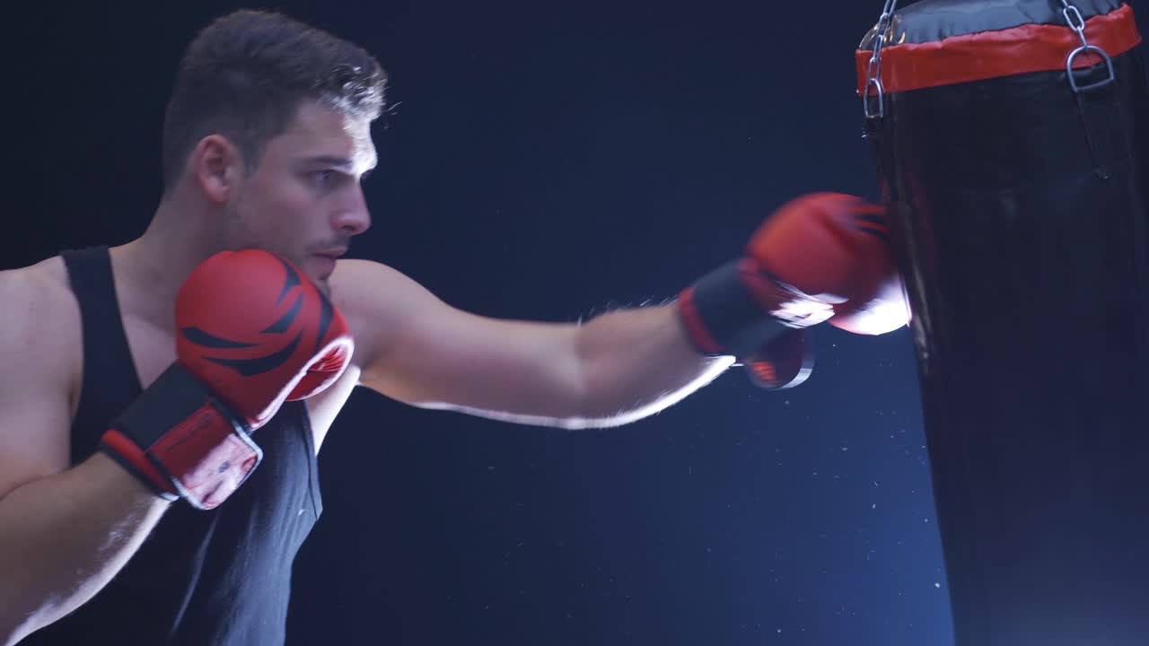 강한 운동선수가 먼지 가득한 체육관에서 박스백을 때립니다. 영화적인 권투 장면입니다.