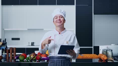 chef asiático sonriendo y señalando a la tableta en su mano mientras está de pie en la cocina de casa